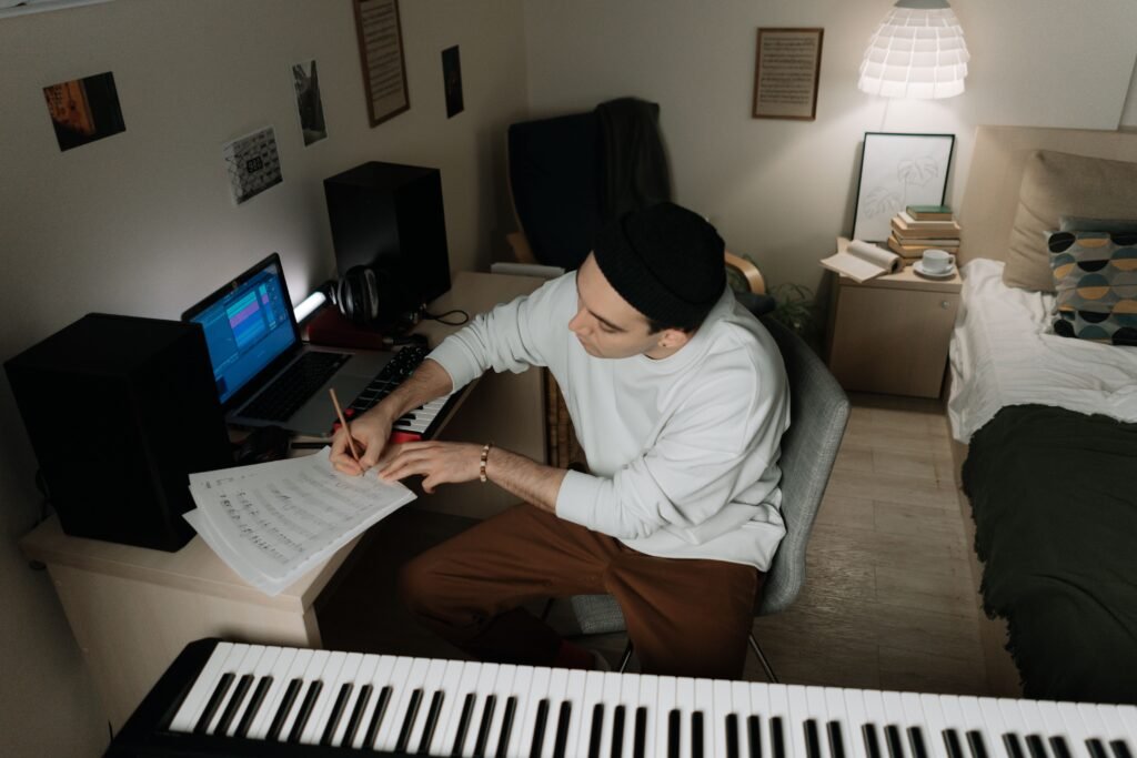 A man in a cozy room writing music notes by his desktop setup with speakers.